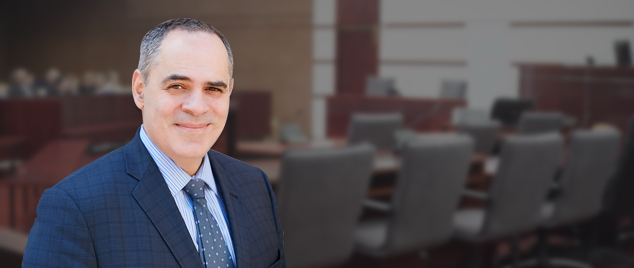 Portrait of Joe Serpa, dressed in a professional blue suit with a patterned tie, standing in the foreground with a blurred courtroom background.