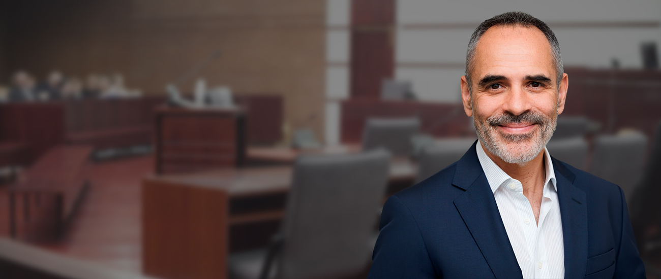 Portrait of Joe Serpa, dressed in a professional suit, standing in the foreground with a blurred courtroom background.